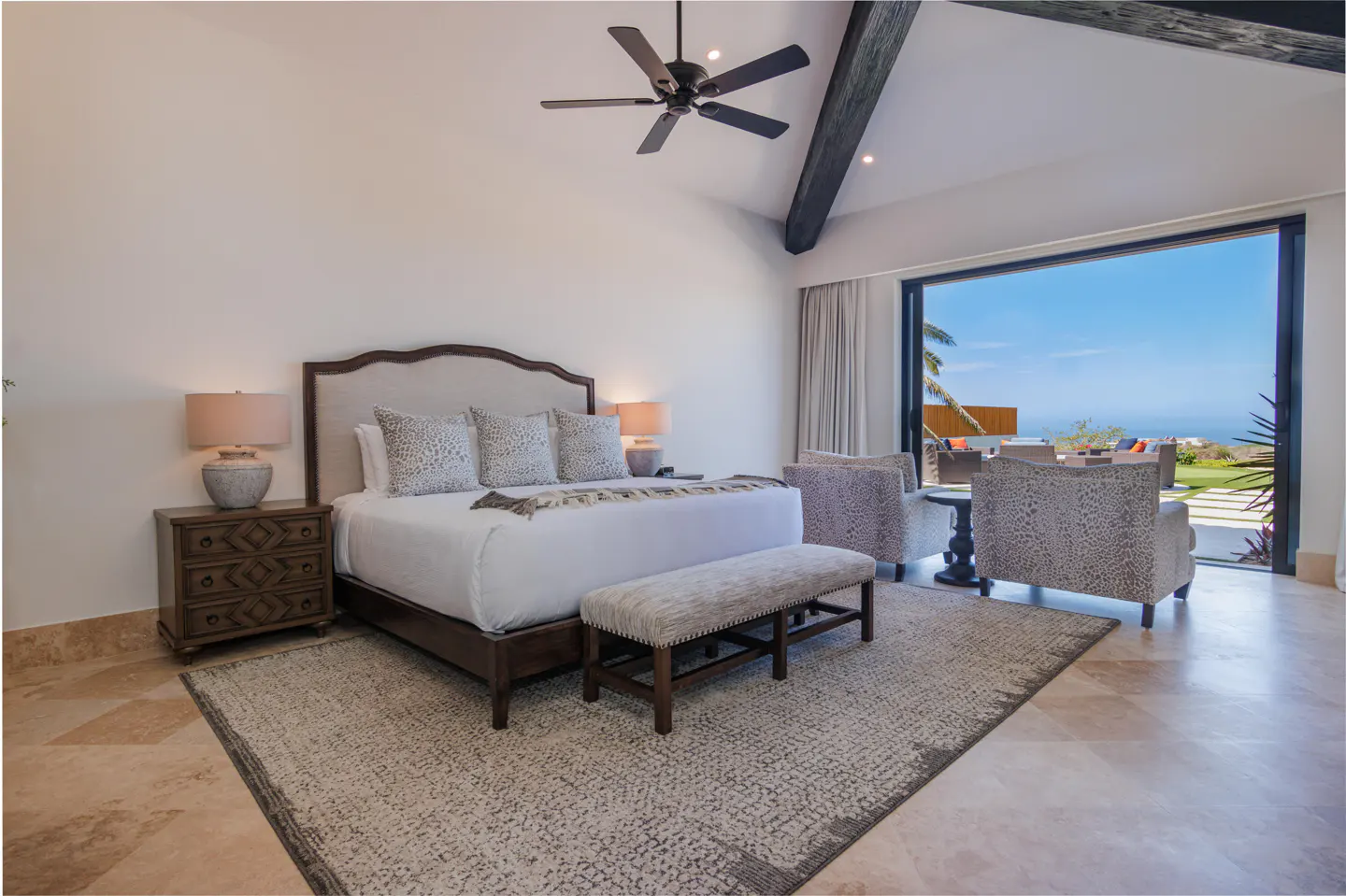 Luxury bedroom with a king bed, patterned rug, and large sliding glass doors opening to a sunny ocean view patio.