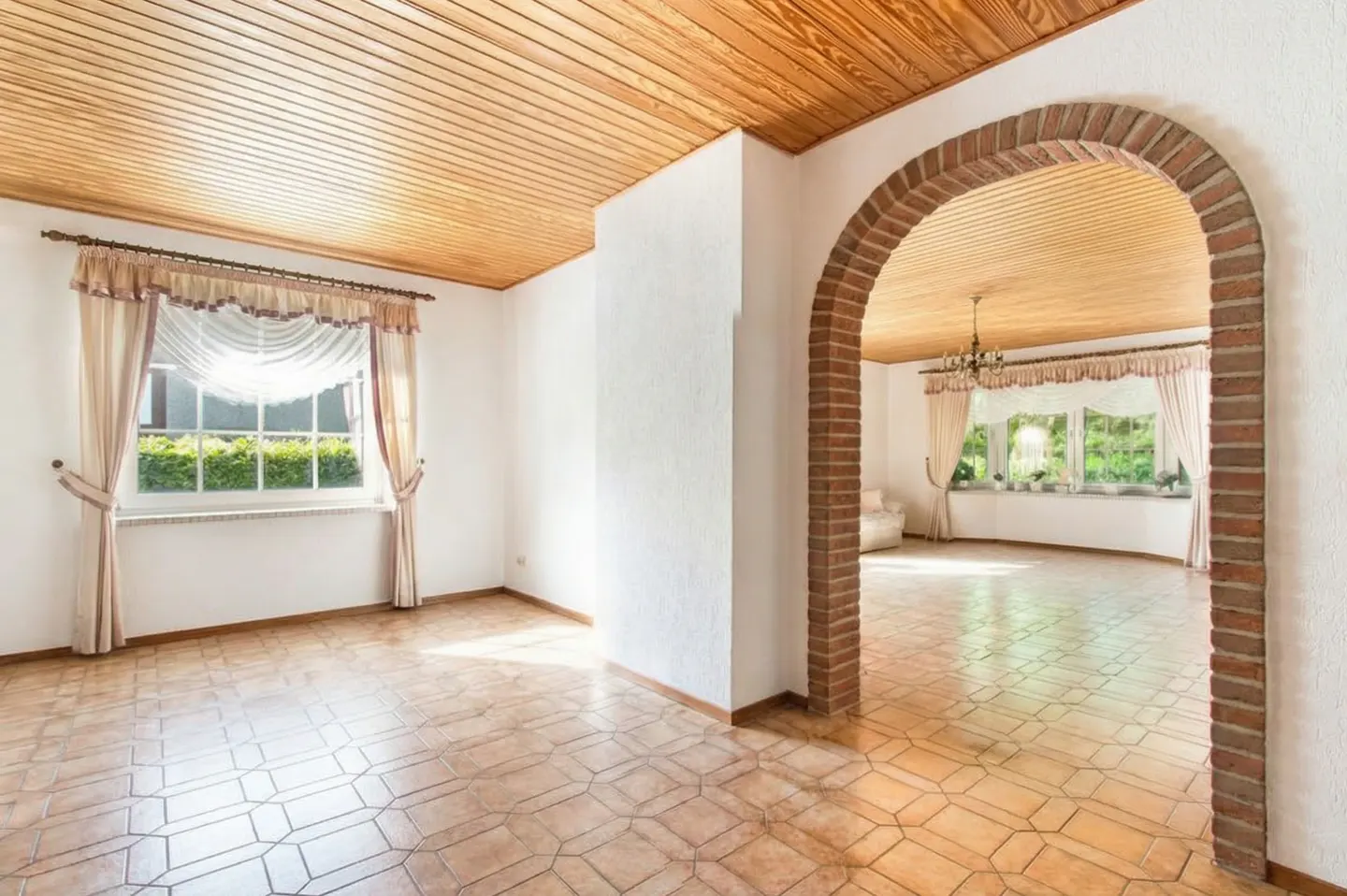 Bright, empty room with a brick archway leading to another room. Wood ceiling, tile floor, and windows with curtains.