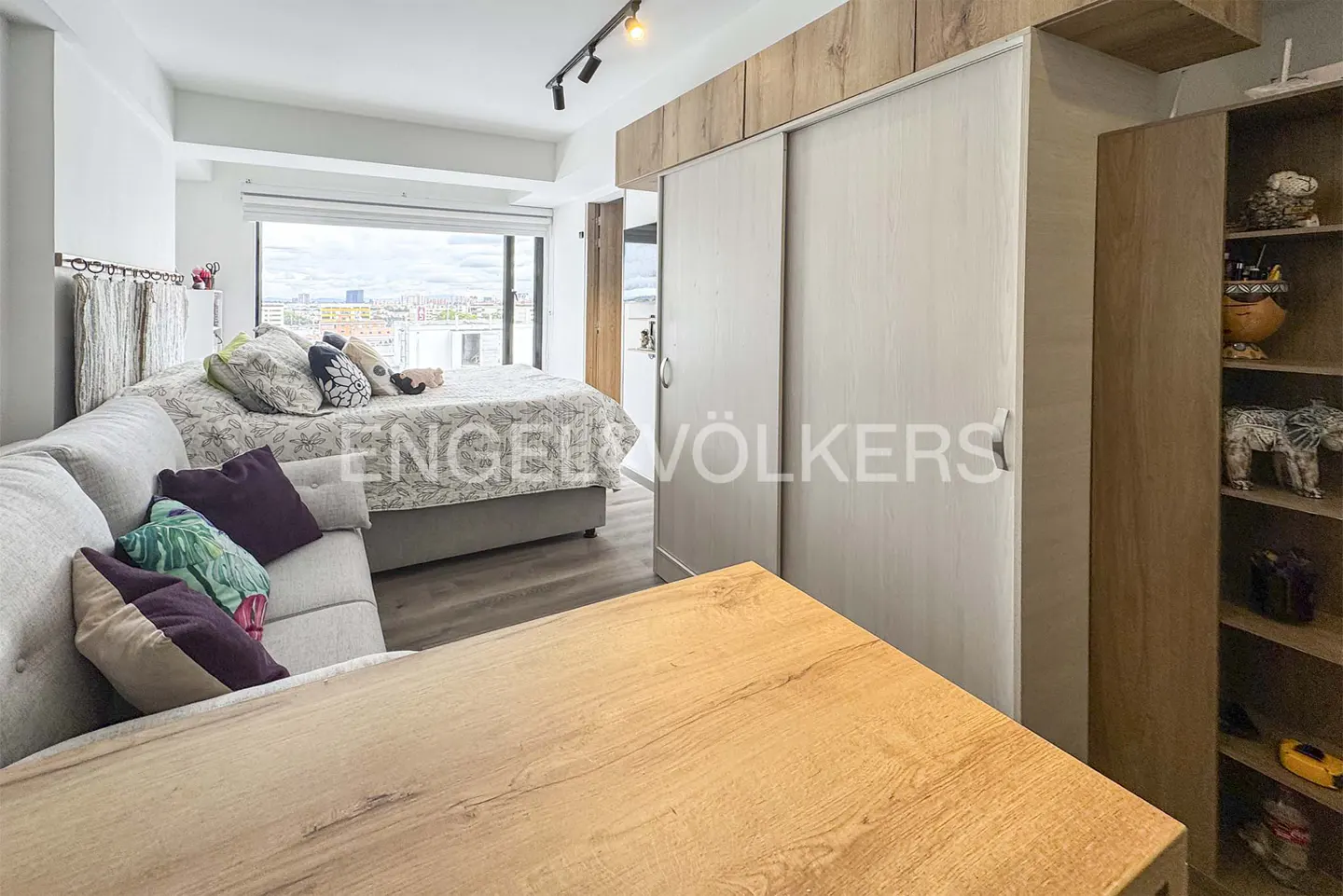Studio apartment with a gray sofa, patterned bed, wood table, and sliding closet doors. A bookshelf is on the right.