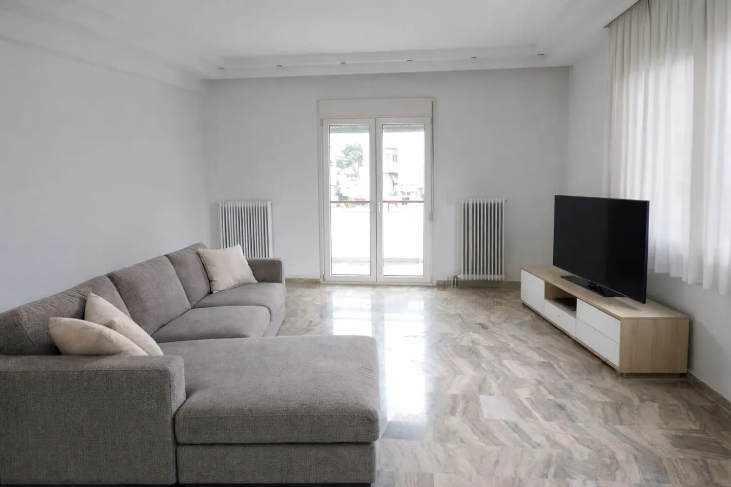 Bright living room with a gray sectional sofa, marble floors, and a TV on a light wood stand. White walls and curtains add to the airy feel.
