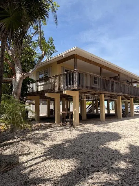 A yellow stilt house with a wooden deck, supported by beige pillars, sits on a gravel lot under a blue sky.