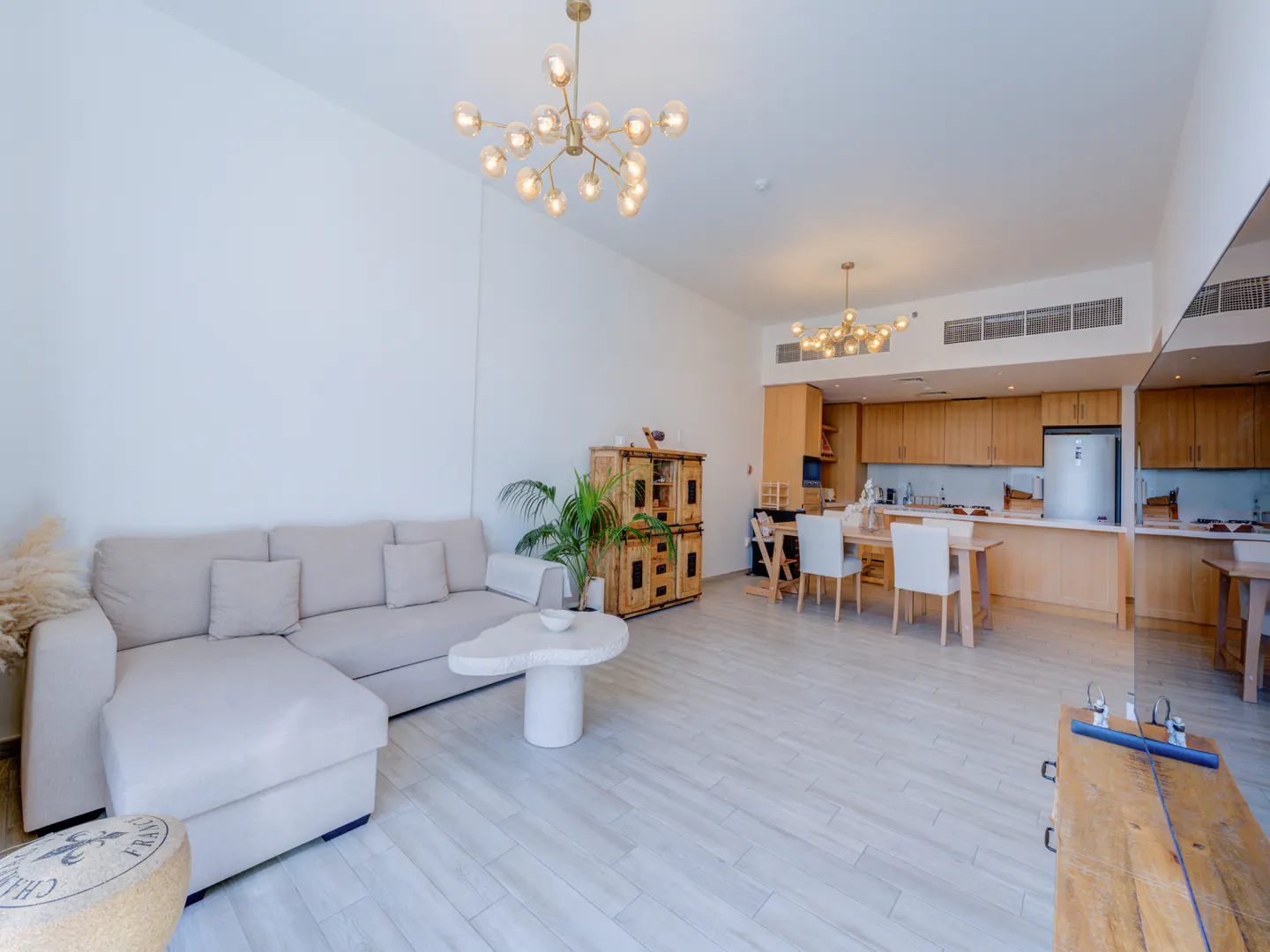 Bright, open-concept apartment with a beige sectional sofa, white walls, and light wood floors. A dining table and kitchen are visible in the background.