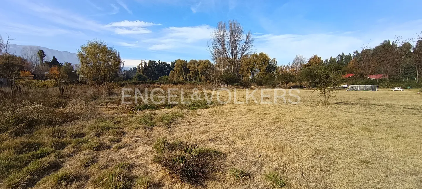 A wide shot of a grassy field with trees, a house, and mountains in the background under a blue sky.