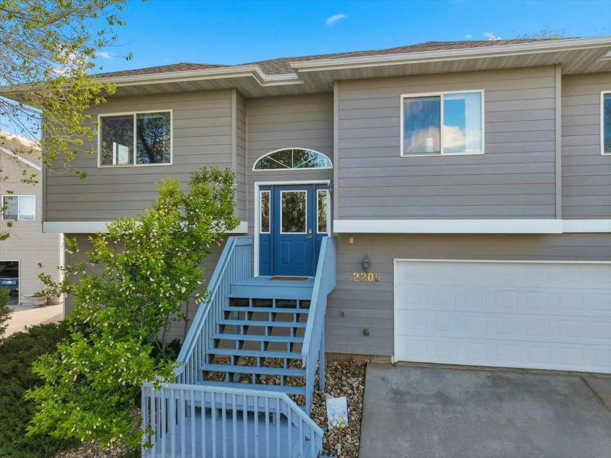 Two-story gray house with blue door and light blue stairs. White garage door on the right. Green tree on the left.