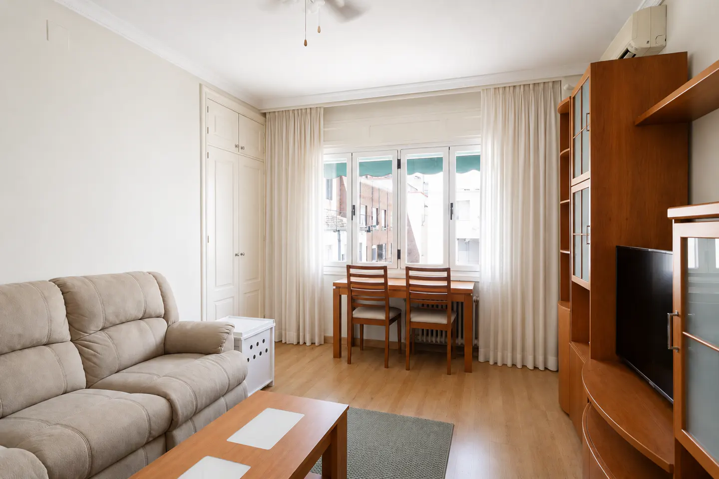 Living room with beige sofa, wood table, and chairs by window with white curtains. A wood entertainment center stands to the right.