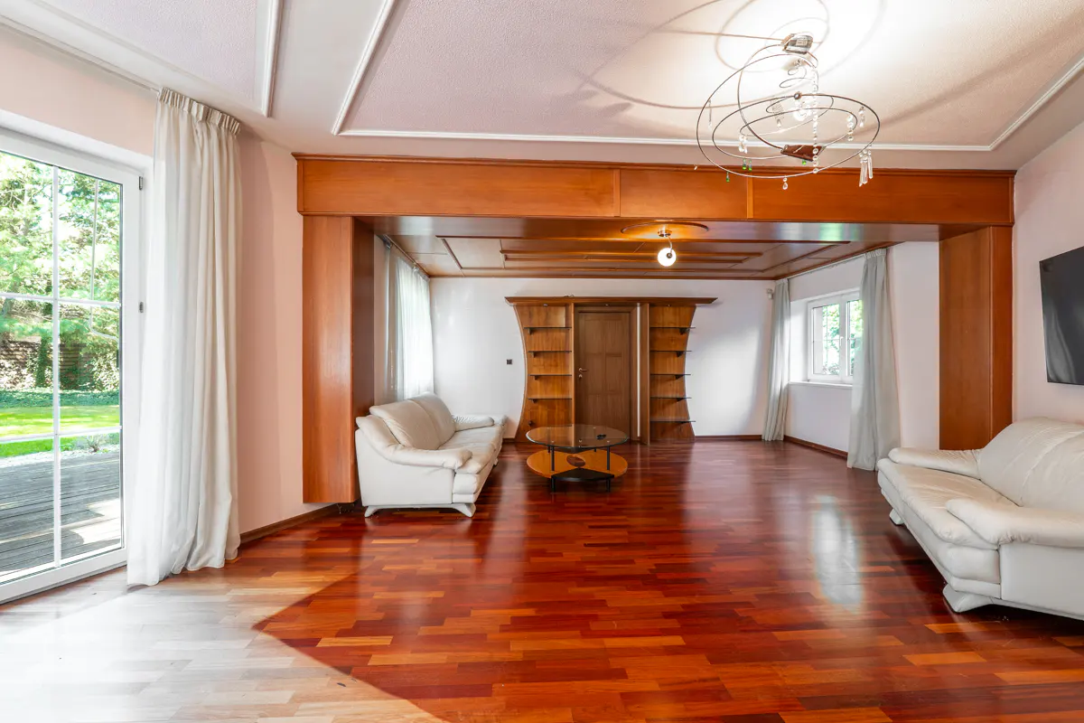 A spacious living room with hardwood floors, white sofas, and a built-in wooden bookshelf with a door in the center. Natural light streams through a large window.