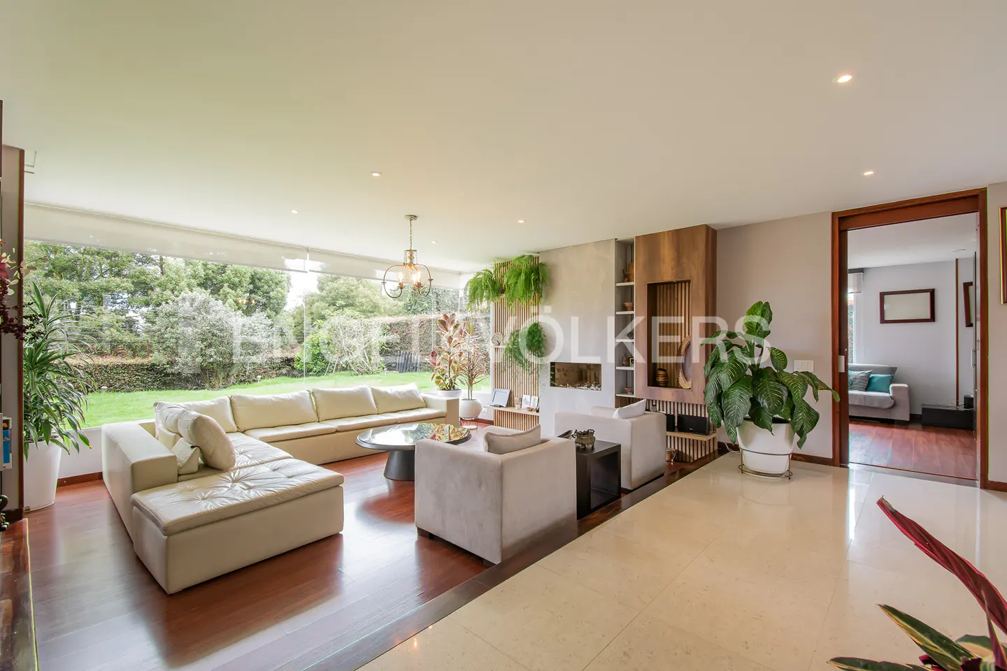 Modern living room with cream sectional, wood floors, fireplace, large windows overlooking a garden, and indoor plants.