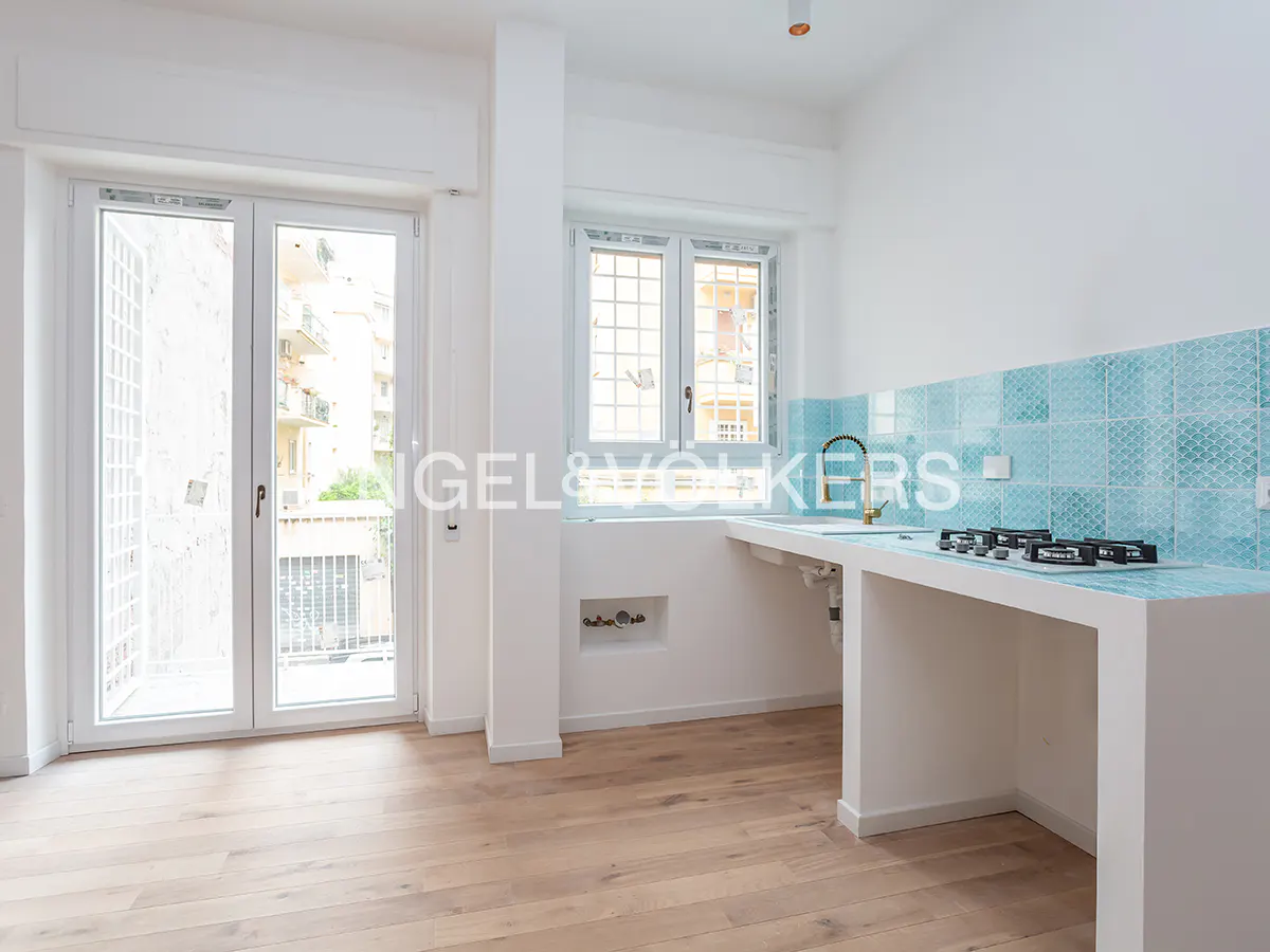 Bright, modern kitchen with light wood floors, white walls, and blue tile backsplash. A window and glass doors let in natural light.