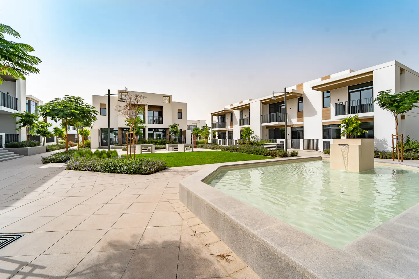 Exterior view of modern white townhouses surrounding a courtyard with a fountain and green space. Balconies and trees add to the serene setting.