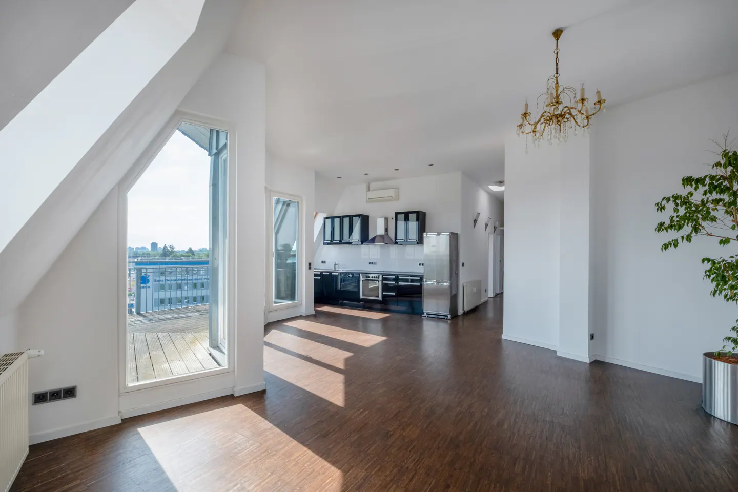 Bright open-plan living area with dark wood floors, a modern black kitchen, a crystal chandelier, and a balcony view.