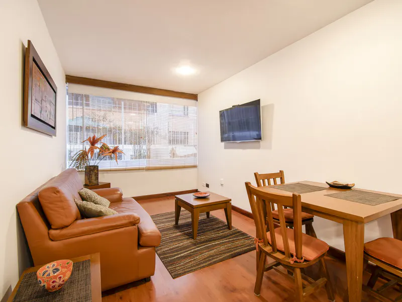 Living room with a tan leather sofa, wooden coffee table on a striped rug, and a dining set against a white wall with TV.