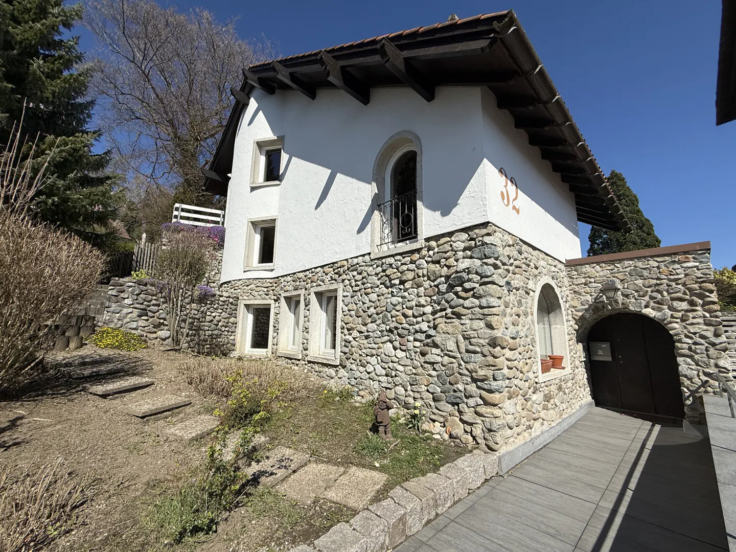 A two-story house with a stone base and white stucco upper level, set on a hillside with a stone walkway.