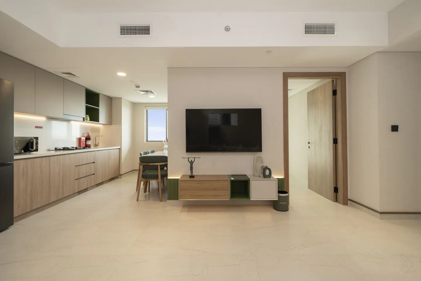 Modern apartment interior with a flat-screen TV above a wooden media console. Kitchen and dining area visible in the background.