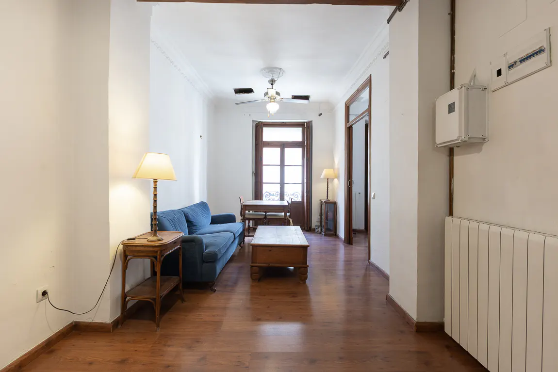 Living room with hardwood floors, white walls, and a blue sofa. A wooden coffee table sits in front of the sofa. A dining table is visible in the background.