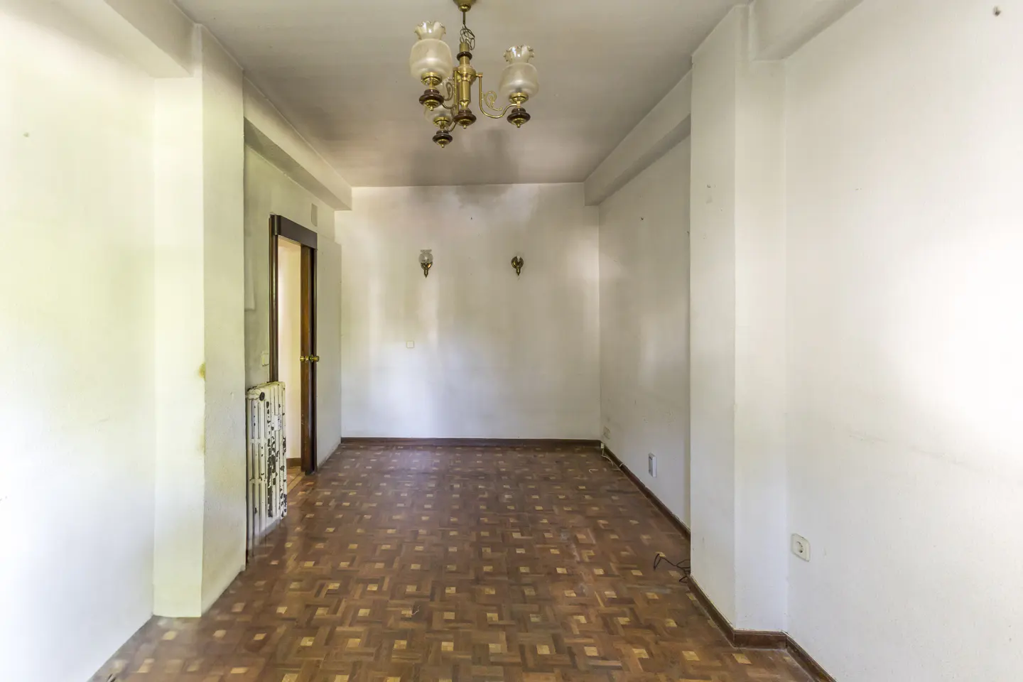 Empty room with white walls, patterned wooden parquet flooring, a vintage chandelier, and a doorway leading to a hallway.