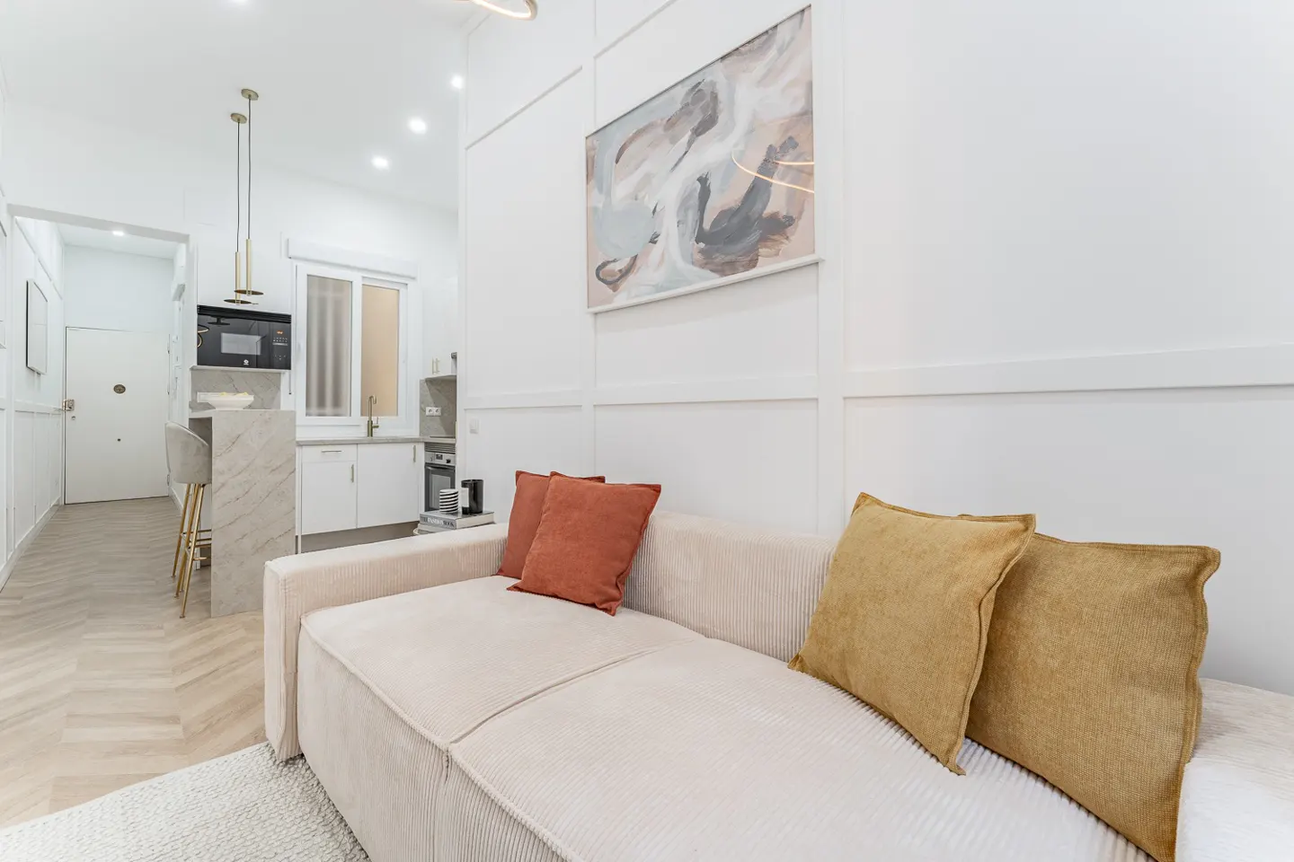 Bright apartment interior with a beige sofa, orange and gold pillows, and an abstract painting on a white paneled wall. A kitchen is visible in the background.