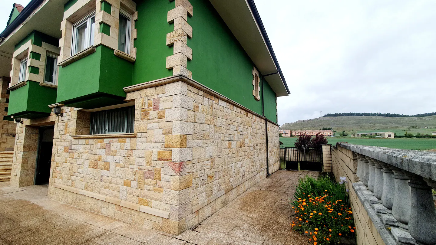 Exterior view of a two-story house with green walls and stone accents. A stone walkway leads to a garden with orange flowers. Rolling hills in the background.