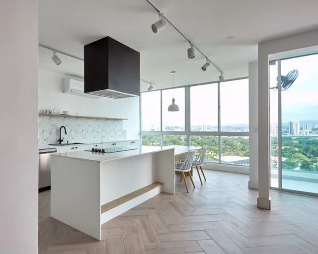 Modern white kitchen with a large island, black range hood, herringbone wood floors, and floor-to-ceiling city view windows.