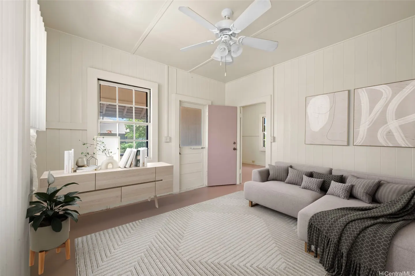 Living room with white walls, a gray sofa with pillows, and a patterned rug. A white ceiling fan hangs above.