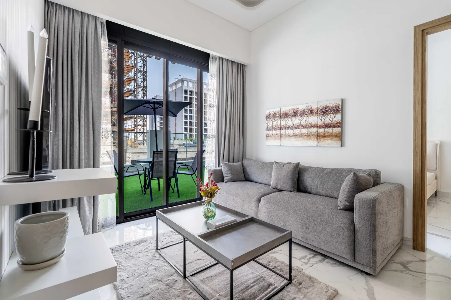 Modern living room with a grey sofa, nested coffee tables, and a balcony view featuring outdoor furniture and a city site.