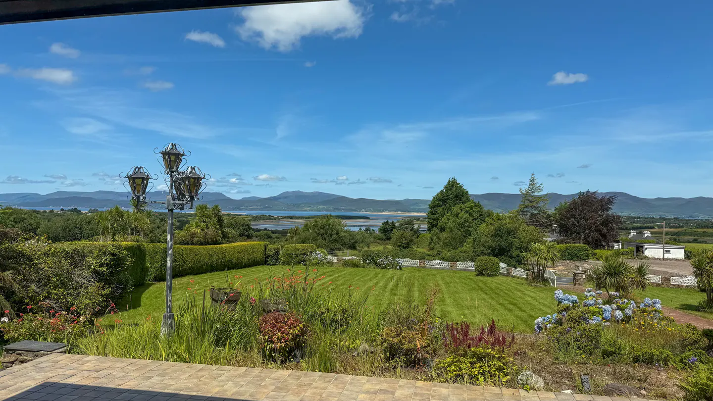 View from a patio overlooking a green lawn, trees, and mountains under a blue sky. A vintage lamp post stands to the left.