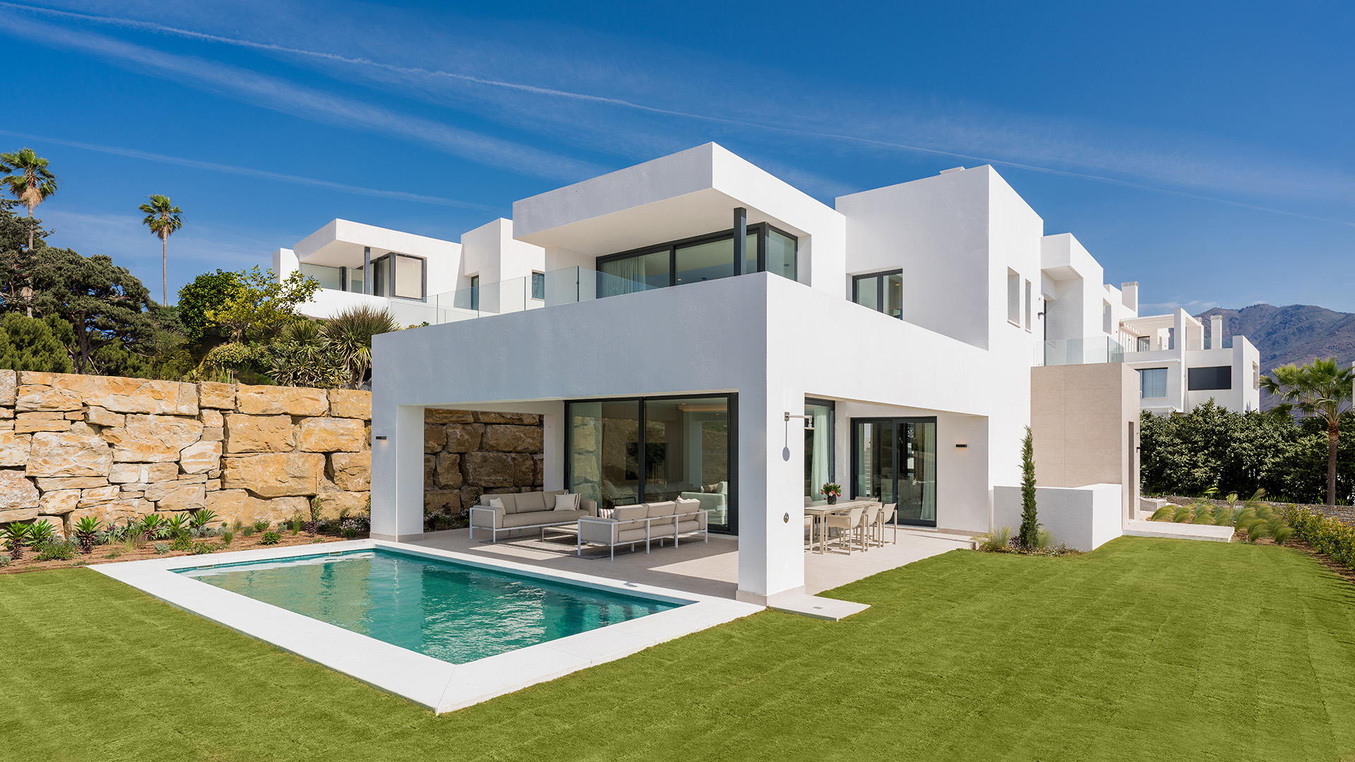 Modern white villa with a swimming pool, patio furniture, and a stone retaining wall against a clear blue sky. [AI-generated text]