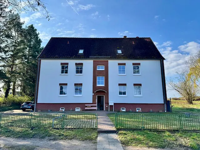 Two-story white building with a dark roof and red brick trim, viewed from across a green fence and grassy yard.