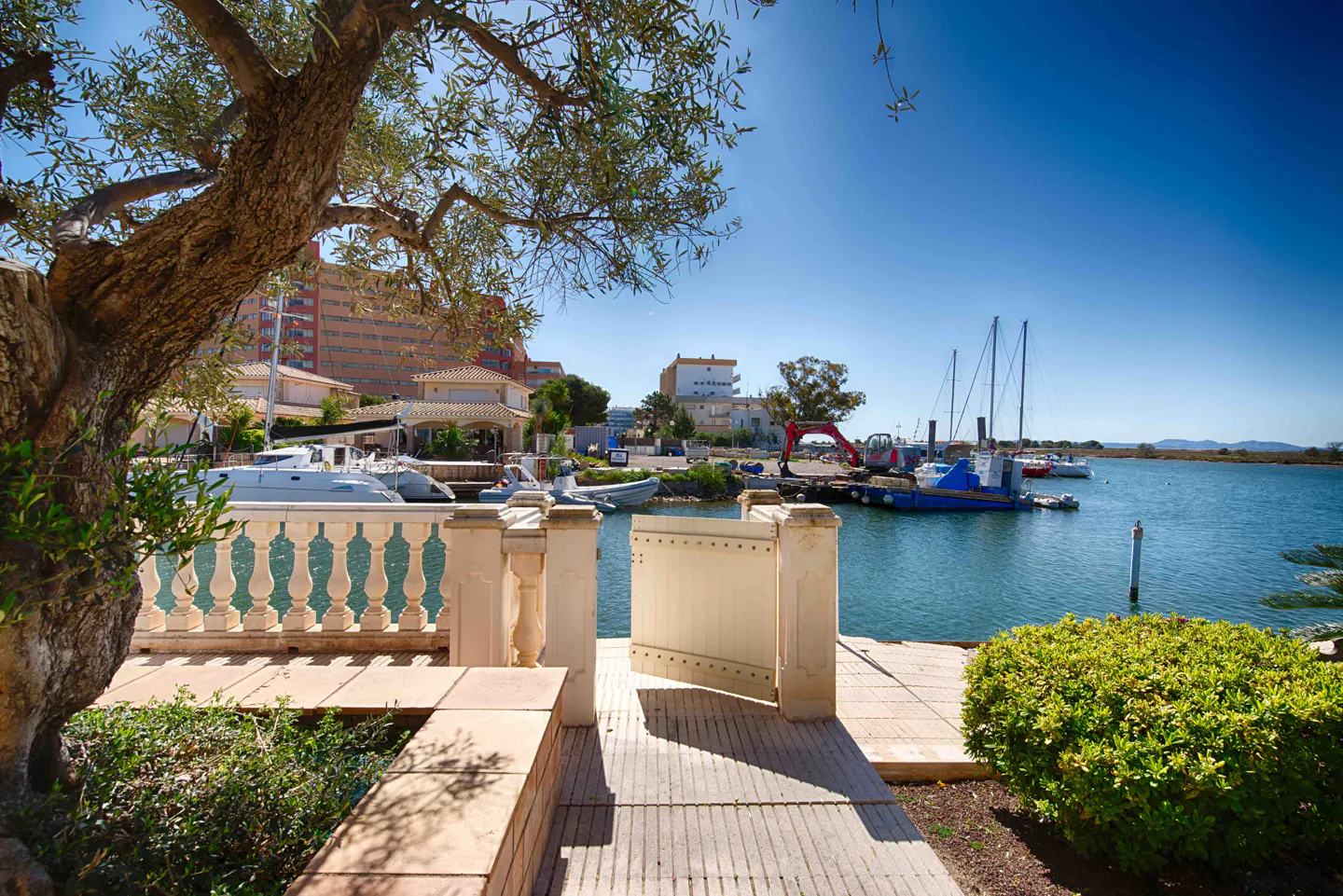 View of a marina from a patio with a white balustrade and gate. Boats are docked in the blue water under a clear sky. A tree and green bushes frame the scene.