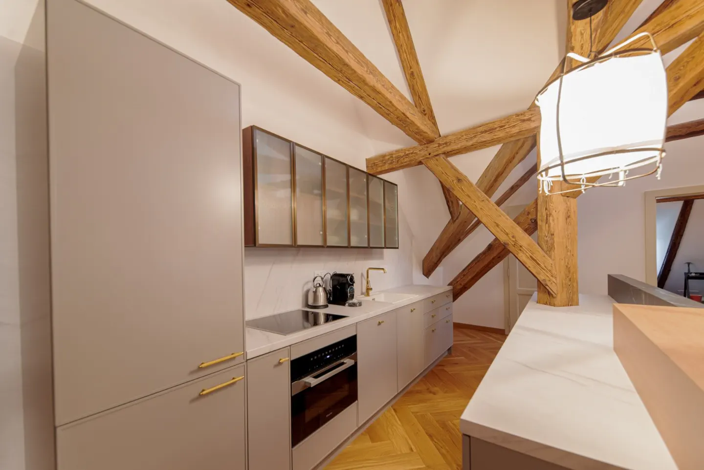 Modern kitchen with light gray cabinets, gold hardware, and white countertops under exposed wooden ceiling beams.