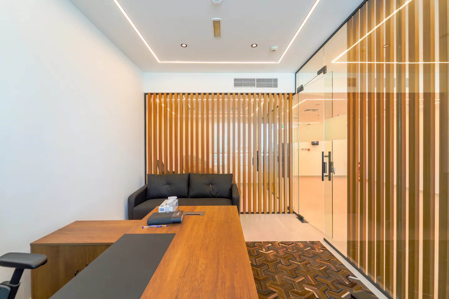 Office interior with a wooden desk, black sofa, and vertical wood slat wall. Glass doors lead to another room.