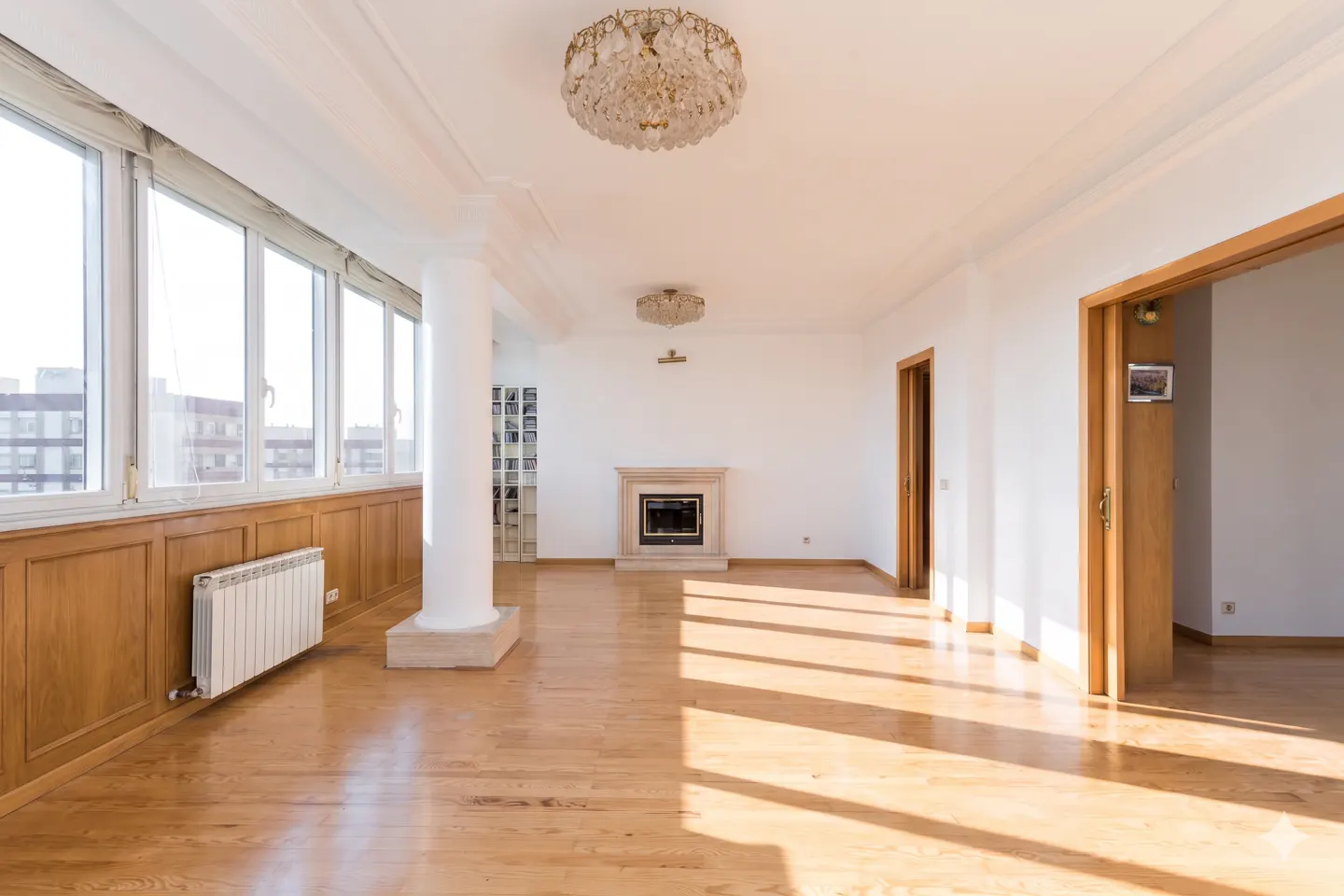 Bright, empty room with wood floors, white walls, and large windows. A fireplace and crystal chandelier add elegance.