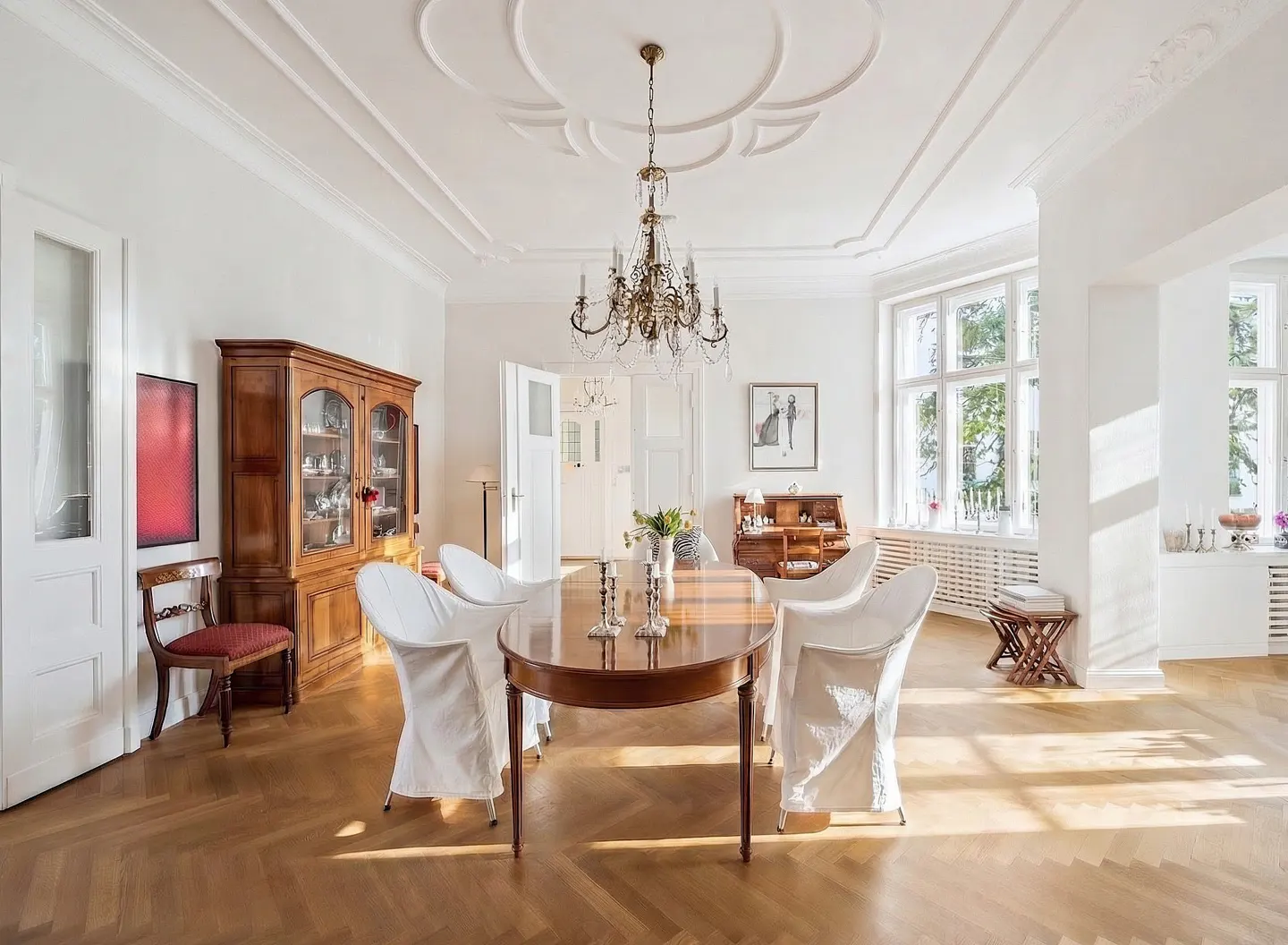 Bright dining room with wood floors, white walls, and large windows. A wood table with white-covered chairs sits under a chandelier.