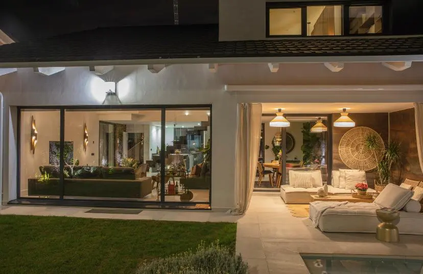Exterior view of a modern home at night with a patio, pool, and open living spaces. Interior lights illuminate the rooms.