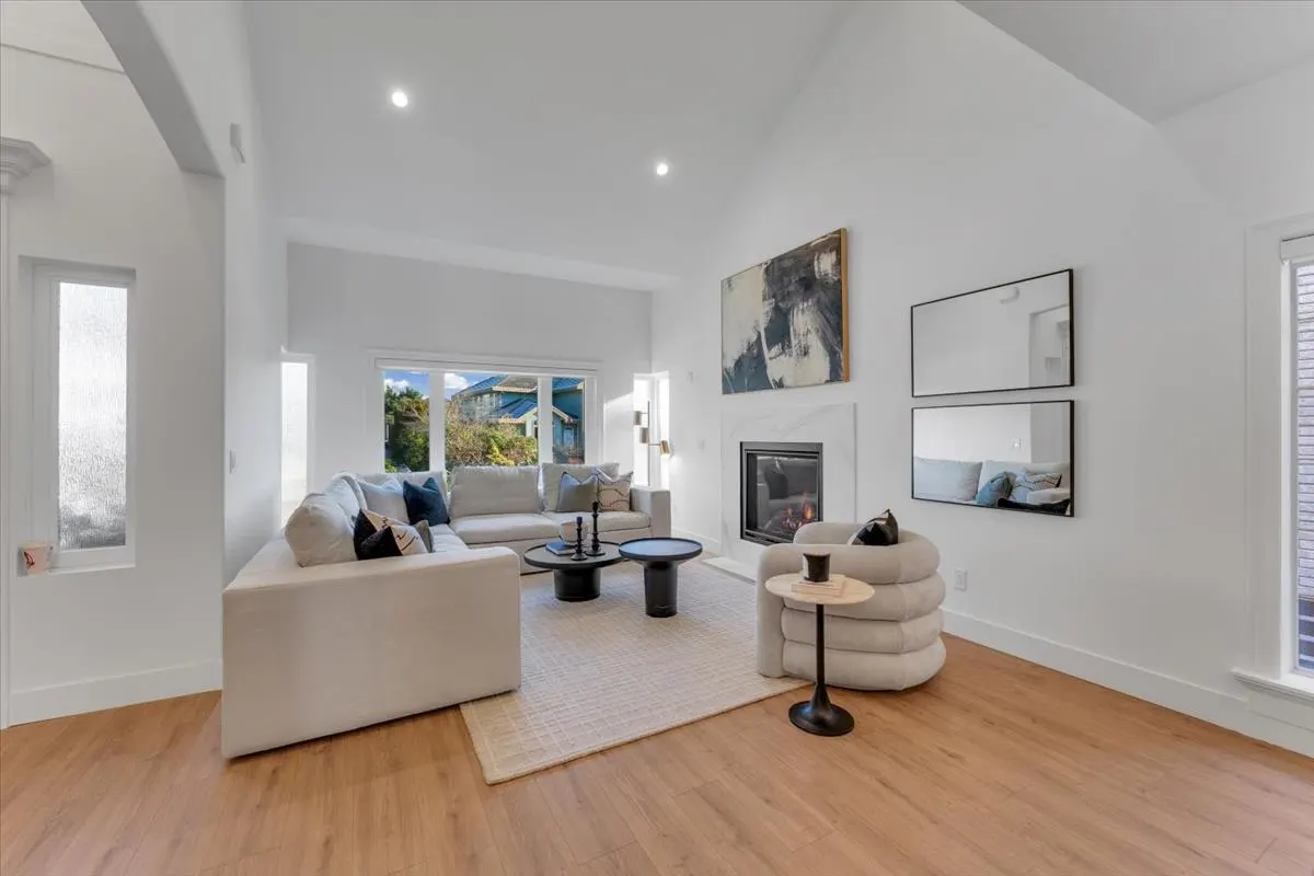 Bright living room with white walls, wood floors, and a fireplace. Two sofas, a chair, and tables sit on a beige rug. Artwork hangs above the fireplace.