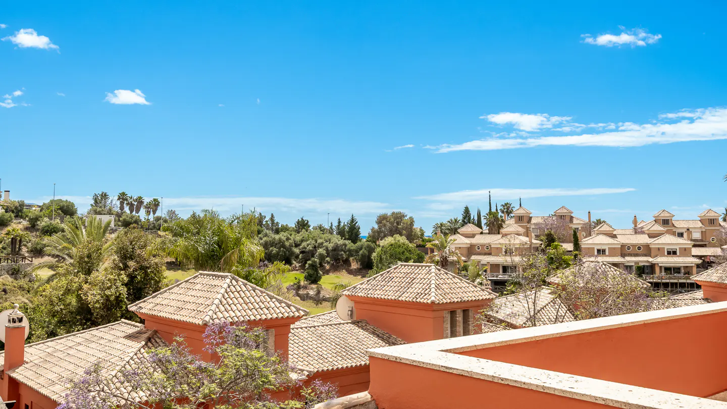 View of terracotta rooftops and buildings under a blue sky, surrounded by green trees.