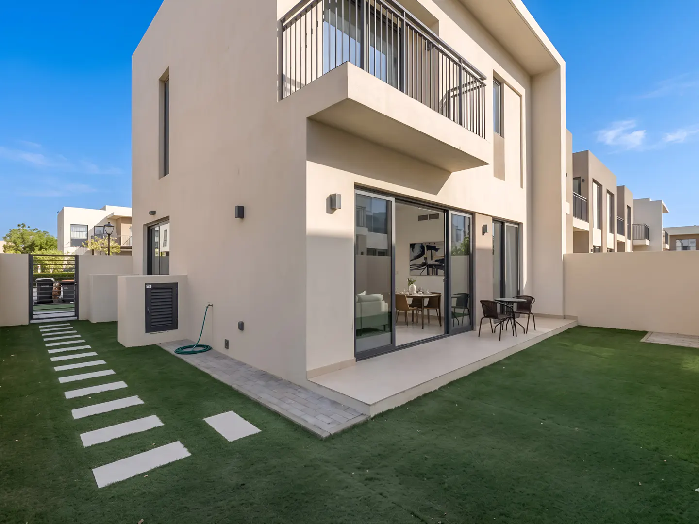 Modern two-story beige townhouse with a private backyard, artificial grass, patio, and balcony under a clear blue sky.