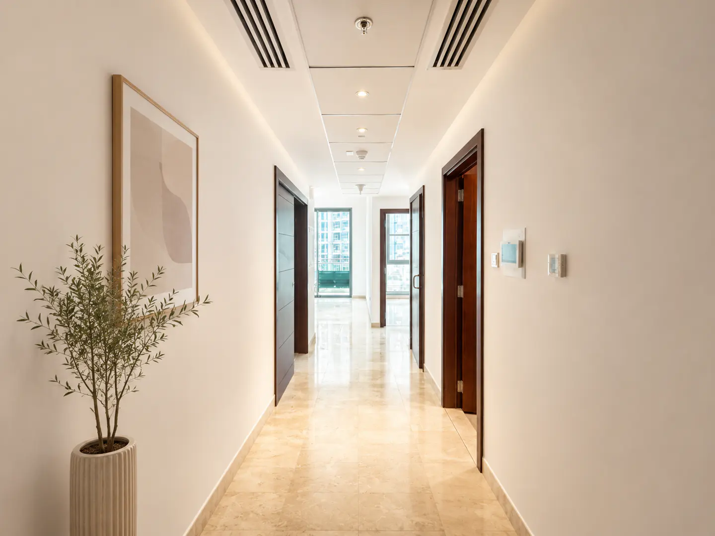 Hallway view with marble floors, white walls, and recessed lighting. A potted plant and framed art accent the left wall. Doors line the hallway.