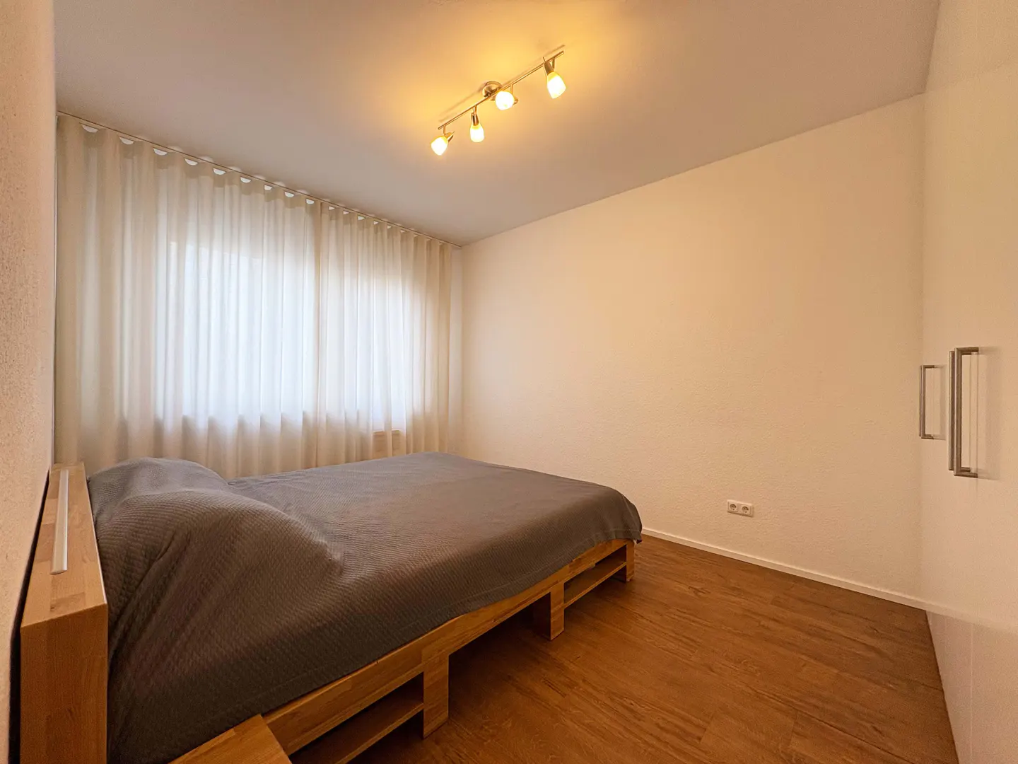 A minimalist bedroom with a wooden pallet bed, grey bedding, white curtains, and warm track lighting over wood floors.