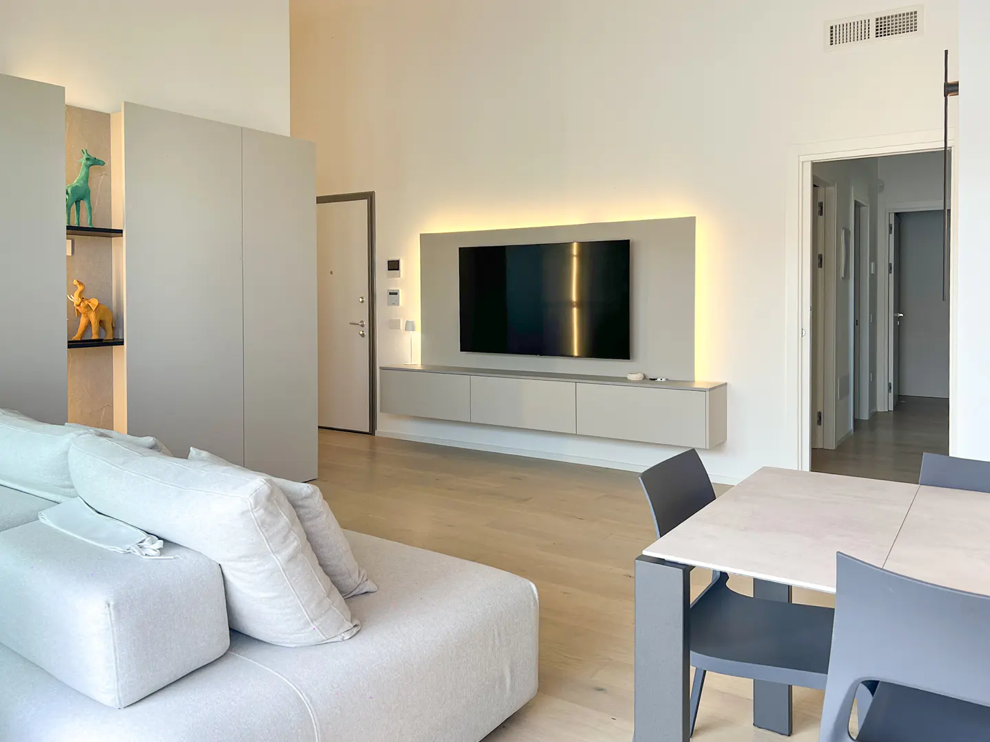 Modern living room with a light gray sofa, wall-mounted TV, floating cabinet, and a dining table with gray chairs.