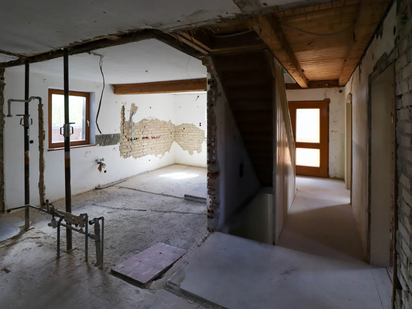 Interior view of a house under renovation, showing exposed brick, plumbing, and a wooden staircase. The walls are white, and the floor is concrete.