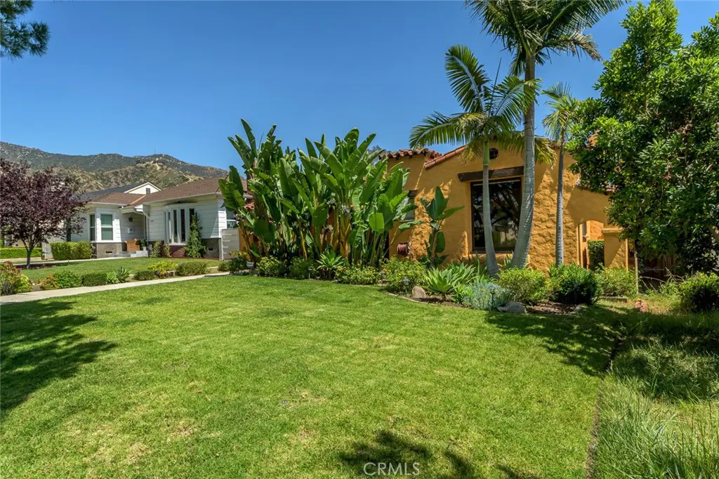 A yellow stucco house with a red tile roof sits on a green lawn with palm trees and other plants. A white house is visible in the background.
