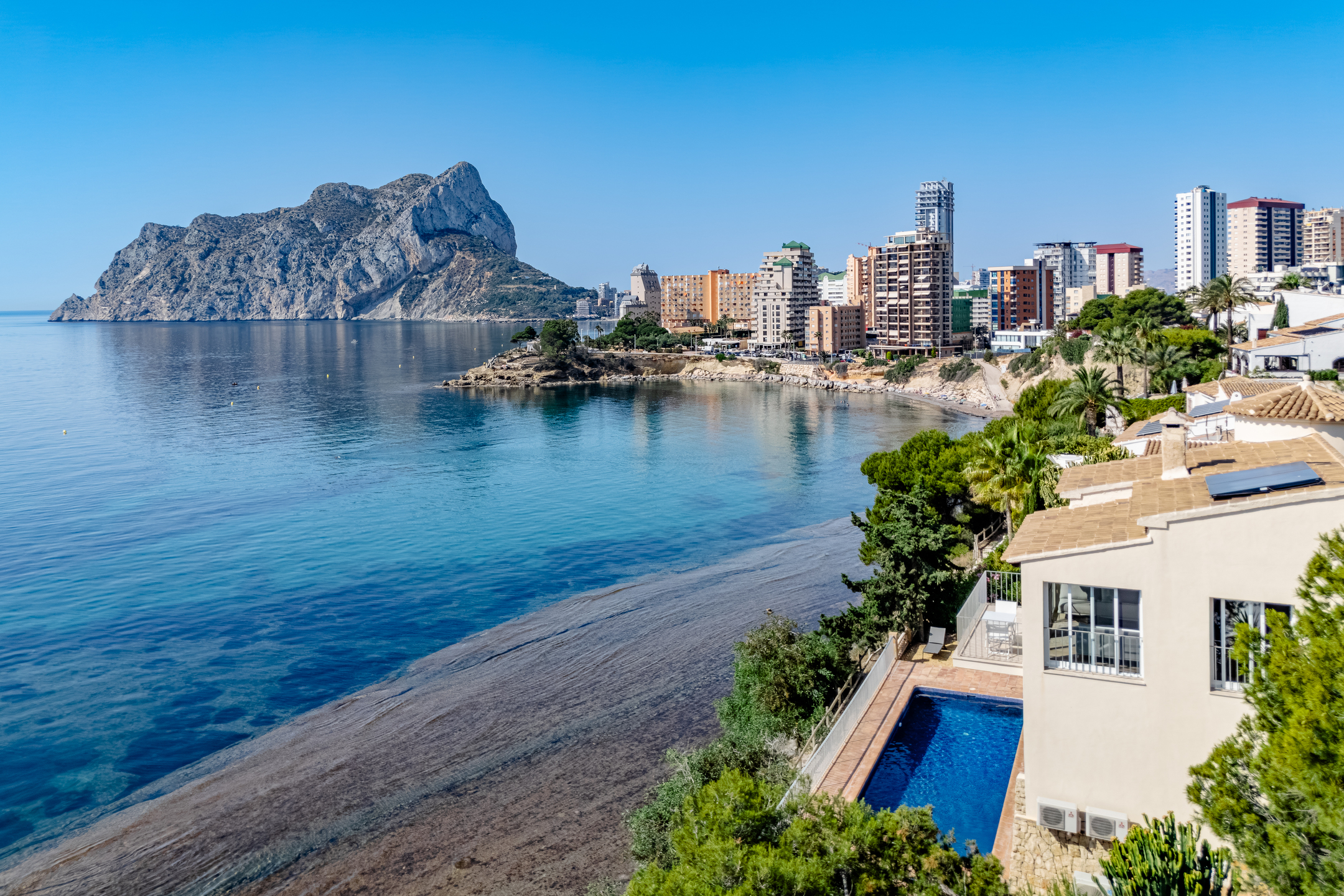 A coastal villa with a private pool overlooking a blue bay, sandy beach, and the Penyal d'Ifach rock in Calpe, Spain. [AI-generated text]