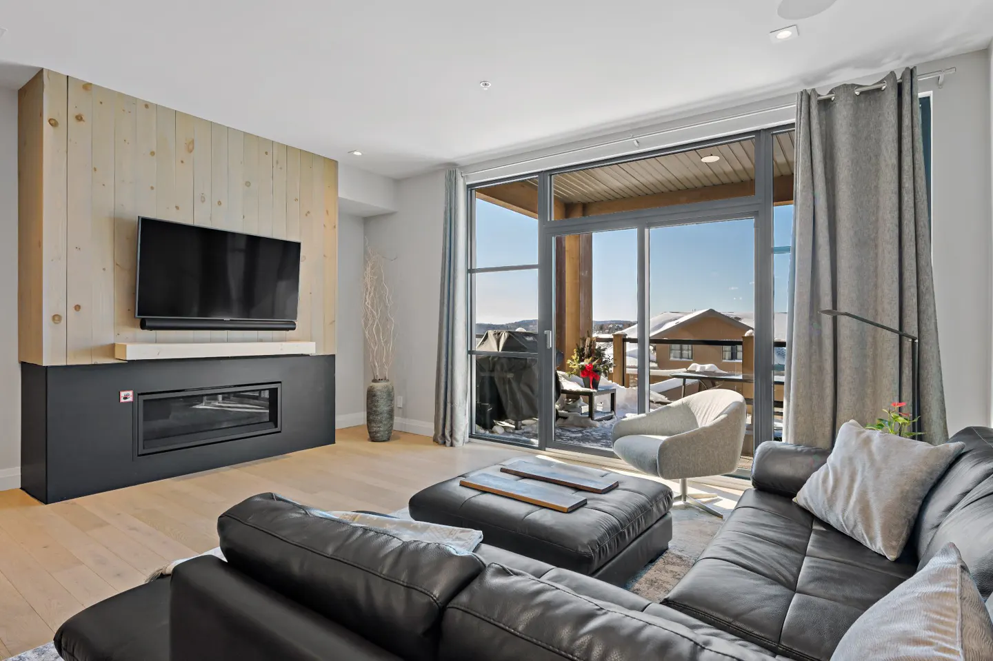 Living room with a black leather couch, fireplace, TV, and a balcony with a snowy view.