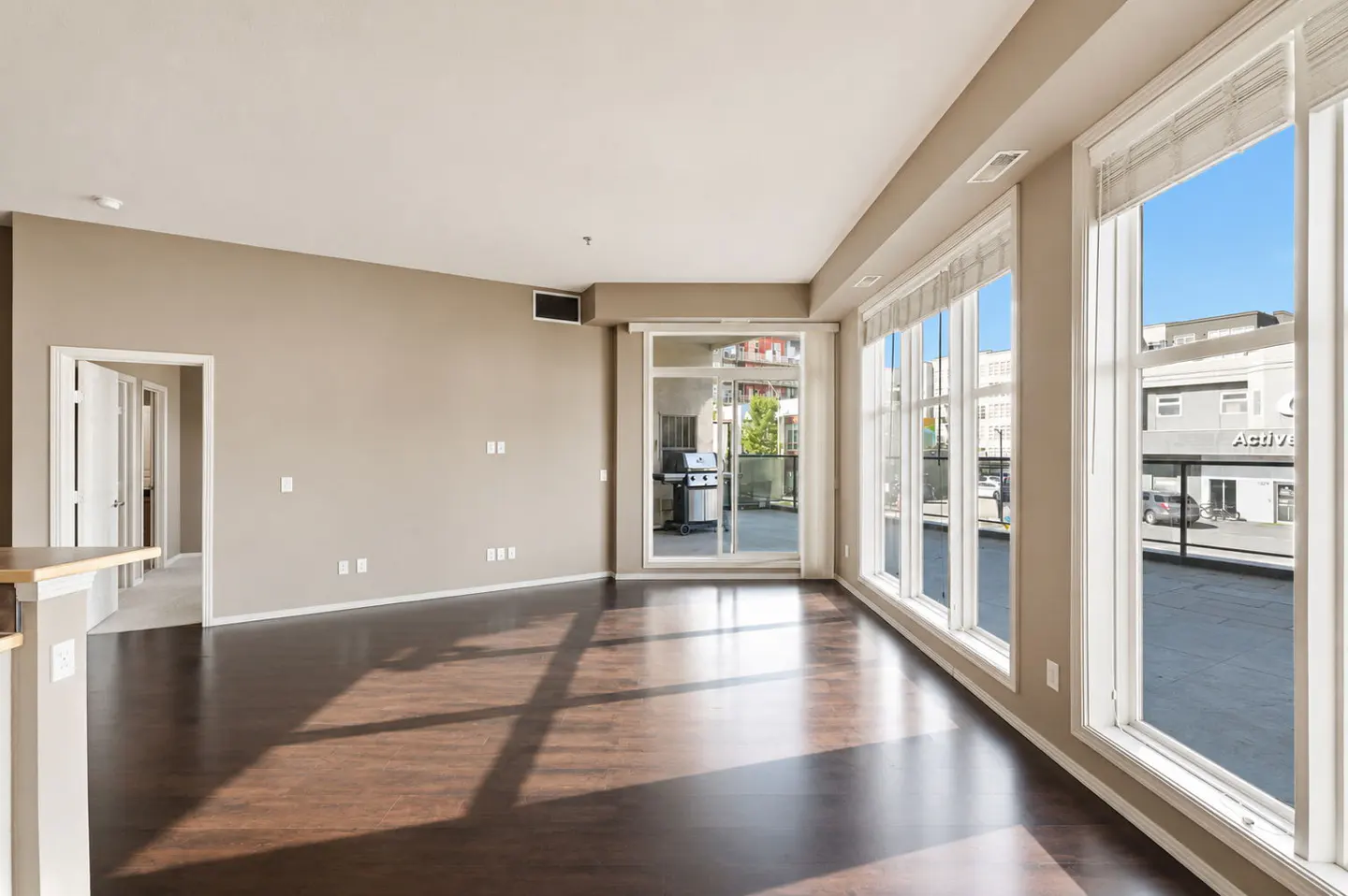 Spacious living room with dark hardwood floors, beige walls, large windows, and a glass door leading to a balcony.