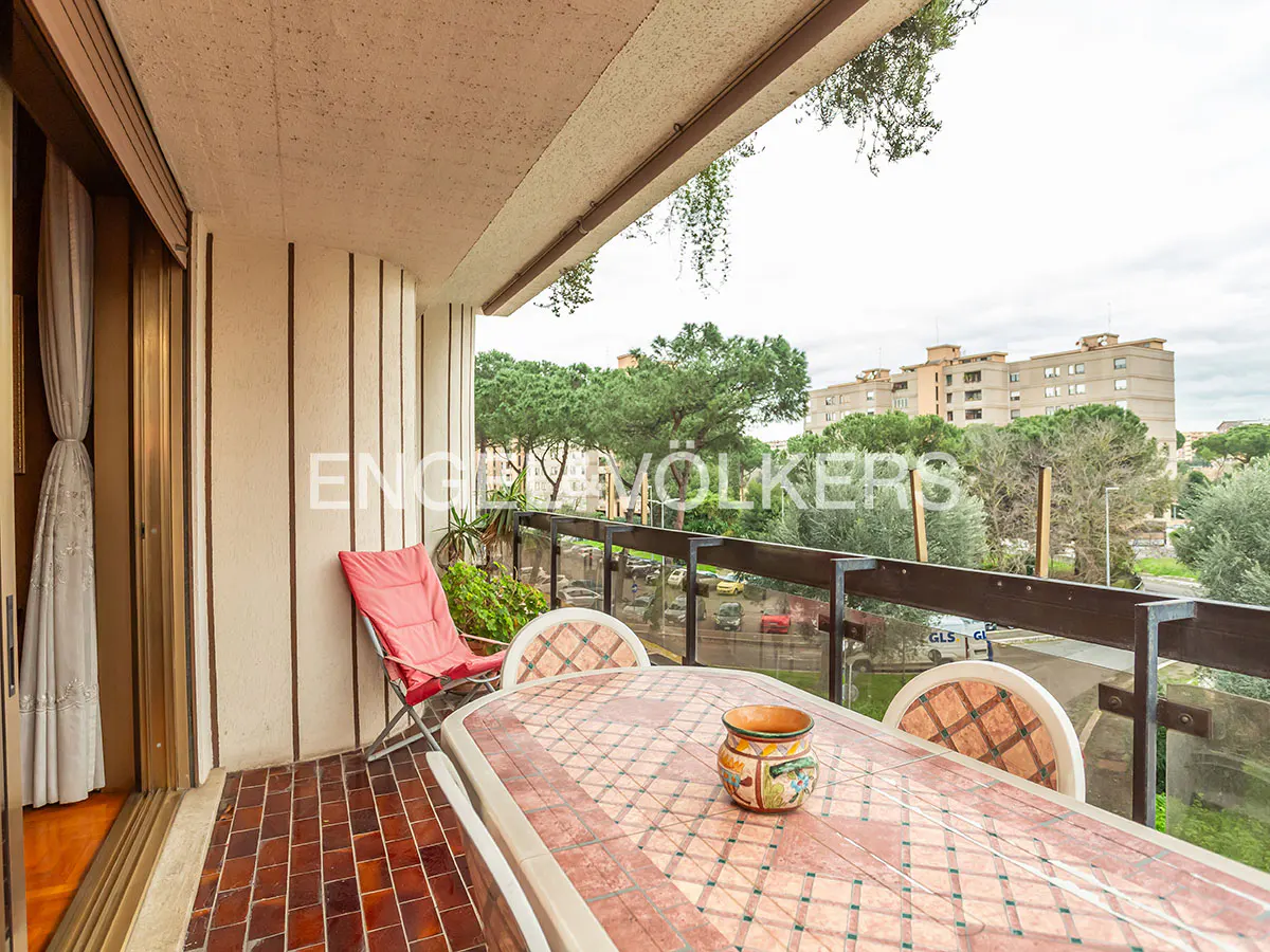 Balcony with red tile floor, table, chairs, and red folding chair. City view with trees and buildings in the background.