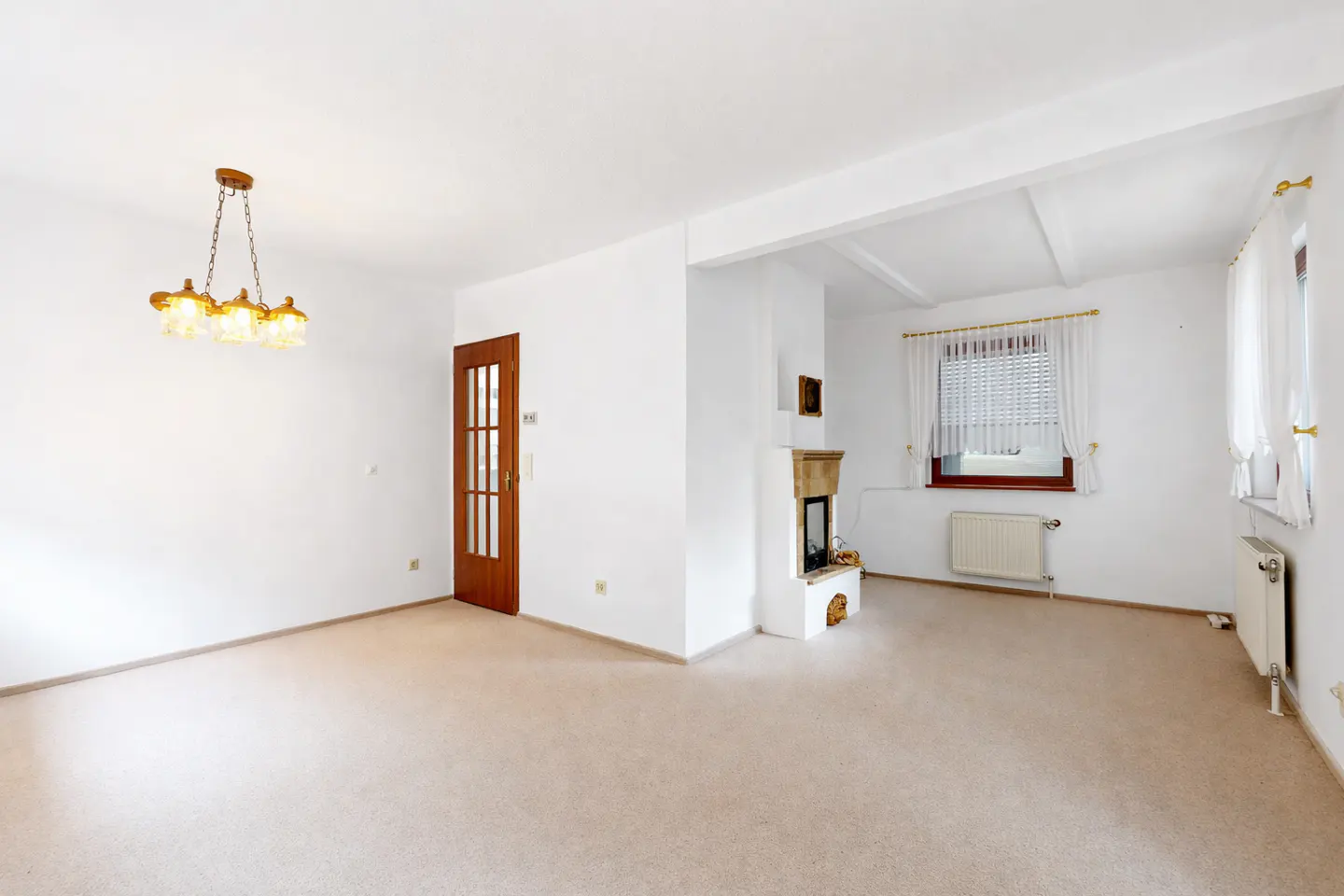 Bright, empty living room with beige carpet, white walls, and a wood door. A chandelier hangs from the ceiling. A fireplace and windows are in the background.