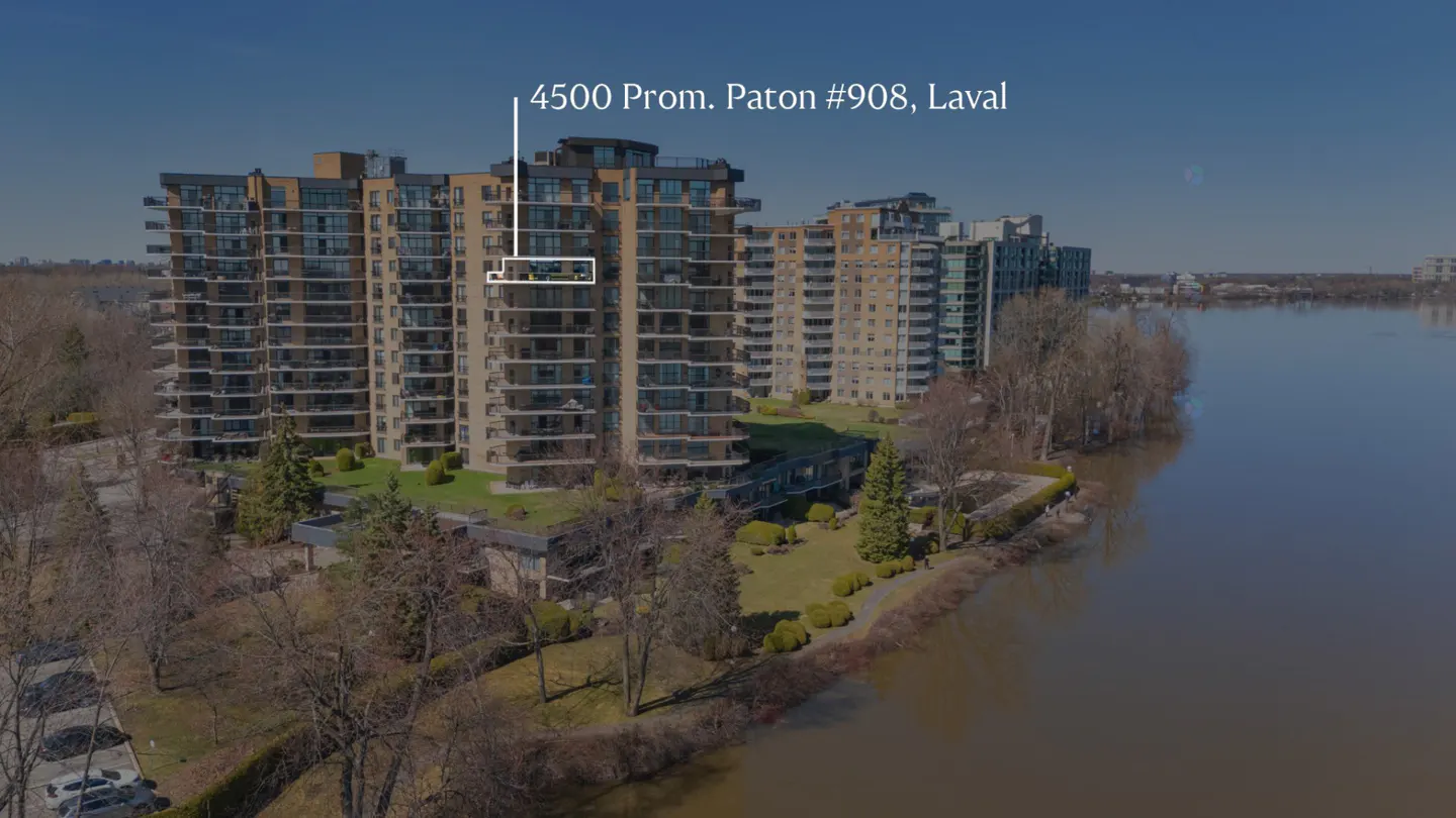 Aerial view of a tall, tan apartment building with balconies, next to a river and trees under a blue sky.