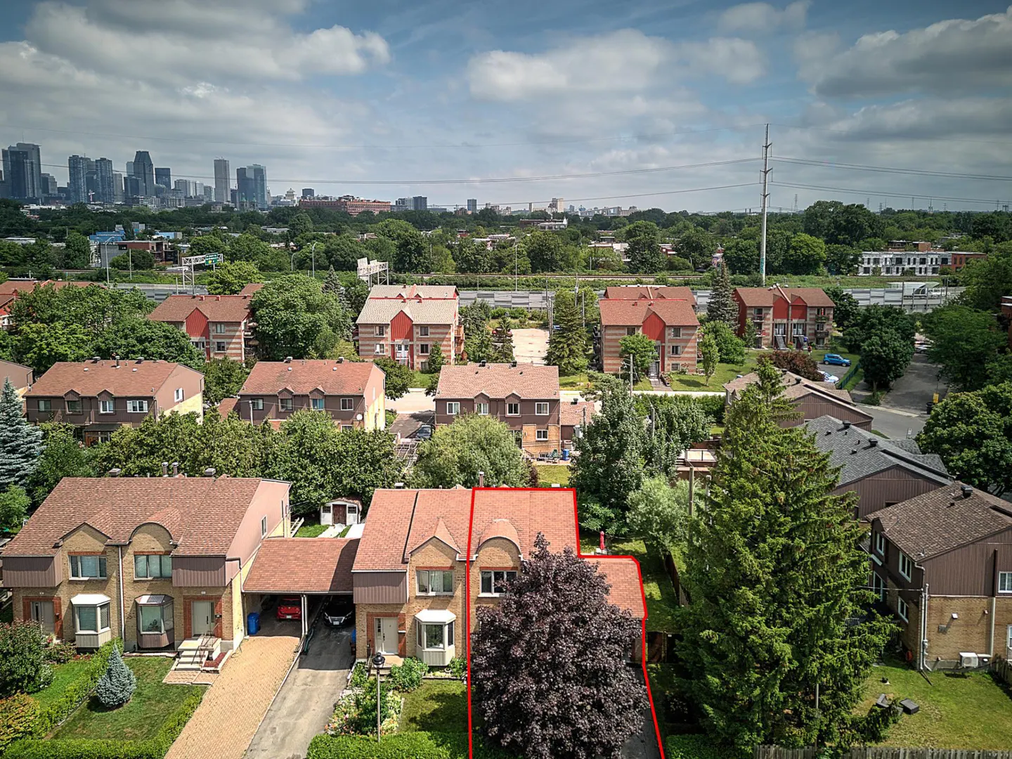 Aerial view of a two-story townhouse with a red outline, brown roof, and green lawn. Other townhouses and a city skyline are in the background.