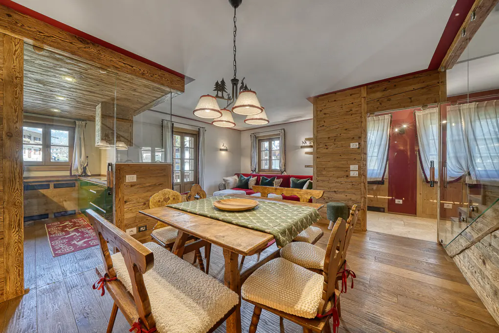 A rustic dining area with a wooden table, chairs with cushions, and a green patterned tablecloth. The room features wood paneling, a unique chandelier, and a glimpse of a cozy living space.