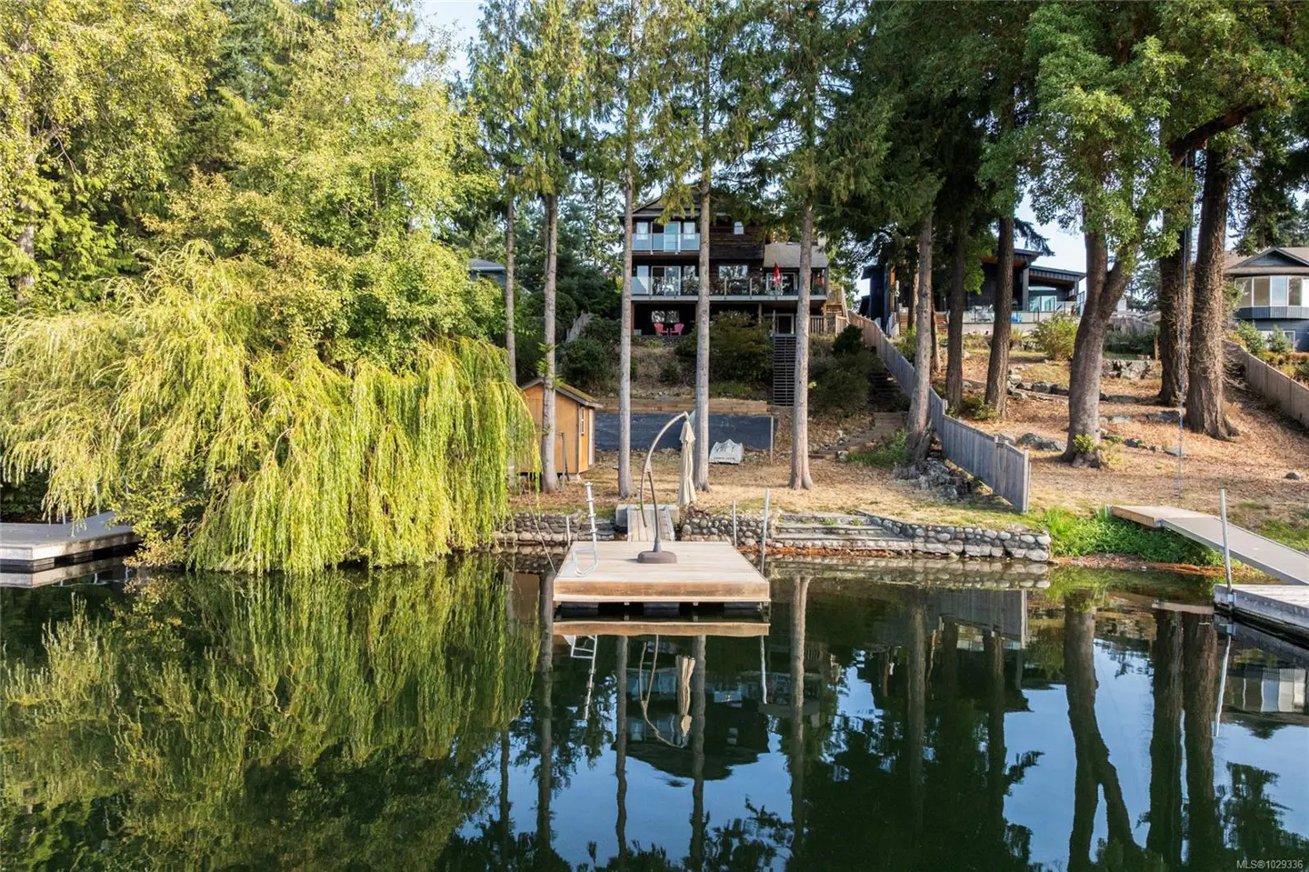 Waterfront home with a dock on a calm lake, surrounded by green trees. The house is two stories with a balcony. Reflections visible in the water.